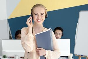 Attractive female hotline operator looking at camera and smiling in modern office. Portrait of helpdesk consultant in headset holding documents, working with her collagues at call centre photo