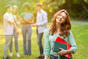 Cute young student girl with workbooks posing outdoors with her classmates on background, resting at campus, enjoying university life, selective focus photo