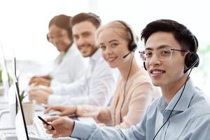 Smiling call center workers sitting in row near their computers at light office, empty space photo