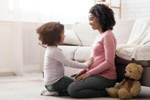 Family Connection. Adorable little daughter touching pregnant mom's belly while playing together on floor in living room, side view with free space photo