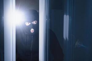 Robbery Concept. Aware masked villain lurking into house through window or glass door, using torch in the dark photo