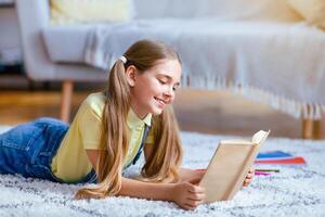 Leisure Concept. Portrait of cute smiling teenage girl lying on the floor carpet at home in living room, holding and reading paper book, enjoying story, copy space. Pupil resting and relaxing photo