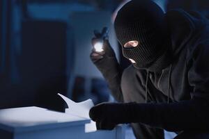 Misappropriation Concept. Masked spy holding flash light and checking papers in office drawer in the dark photo