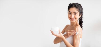 Indian woman with a cheerful expression applies moisturizer to her arms while holding a skincare container. She is in a bright indoor setting, focusing on pampering and self-care, copy space photo