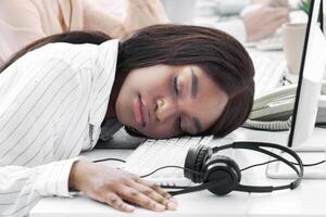 Tired hotline consultant sleeping at her desk, feeling exhausted or stressed after communication with customers. Overworked tech support manager and her colleagues at call centre office photo