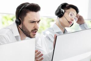 Frustrated helpdesk operator dealing with client's problem in call centre. Millennial tech support agent and his coworker having hard time solving customer's complaint at office photo