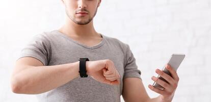 Modern technologies for sport. Guy with fitness tracker on his hand and smartphone, checks activity at light background at home photo