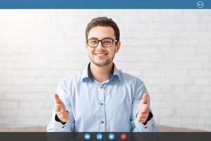 Online job interview. Smiling guy in glasses makes gestures with hands on webcam in interior, free space photo