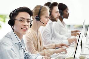 Asian call centre manager smiling and looking at camera in light office, space for text. Helpdesk operator in headset with his colleagues providing technical support services to clients photo