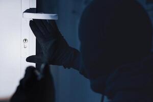 Break-In Concept. Back view of unrecognizable criminal in black balaclava entering victim's house, using torch on lock photo