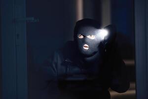 Shadowing Concept. Criminal wearing black balaclava watching you through window or door, using flashlight torch photo