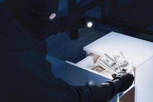 Masked burglar stealing money from drawer, using flashlight, closeup portrait over the shoulder, back view photo