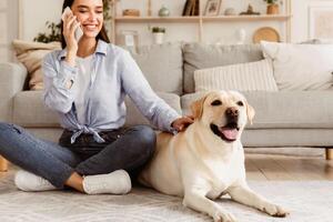 Portrait of beautiful young woman stroking dog and speaking with her friend or relatives using mobile phone at home photo