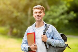 Handsome University Student Guy With Backpack And Workbooks Posing Outdoors At Campus Cortyard, Smiling At Camera, Free Space photo