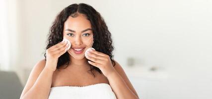 A woman with curly hair smiles while performing a skincare routine, gently using cotton pads on her face. The soft lighting highlights her relaxed expression, emphasizing self-care and rejuvenation. photo