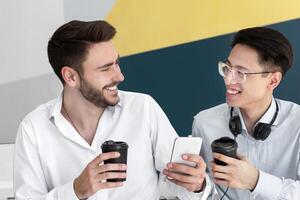 Male helpdesk agents browsing internet on smartphone during coffee break at modern call center, copy space. Positive customer support operators having friendly conversation and hot drink at office photo