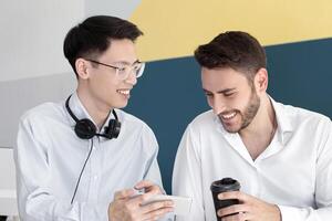 Male helpdesk operators with hot takeout coffee browsing web on smartphone during break at call center, empty space. Tech support consultants chatting and watching videos on mobile device at office photo