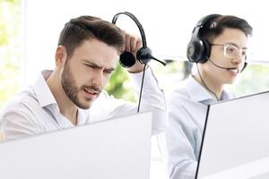 Stressed call center operator listening to client's complaint at contemporary office. Hotline consultant having difficulty dealing with customer's request, feeling stressed and irritated at workplace photo