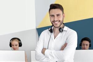 Portrait of positive male customer support representative and his colleagues at contemporary call centre. Happy hotline operator with headset looking and camera and smiling at office photo