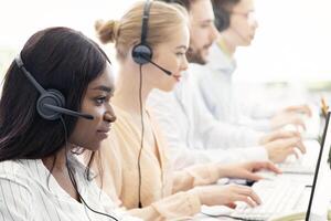 Focused call centre operators with headphones working with computers at open space office, blank space. Multinational team of hotline managers providing telemarketing services to customers photo