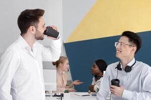 Male hotline consultants drinking coffee during their break at call center. Technical support agents with hot beverages communicating with each other at modern open space office photo