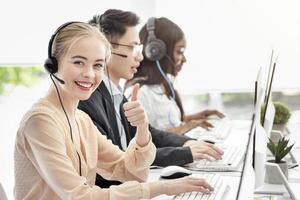 Happy female call centre consultant showing thumb up gesture and her colleages at modern office. Positive helpdesk operator working in telemarketing or in customer support service photo
