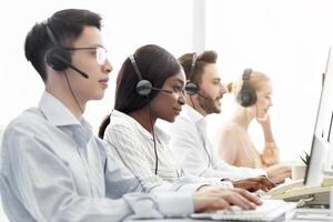 Line of call centre representatives with headsets working on computers at open space office. Group of hotline operators managing customer's claims online, space for text photo