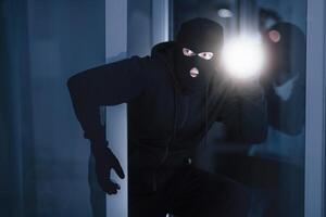 Time For Crime. Cautious burglar looking and peeping into building through window or glass door with torch photo