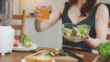 A woman holding a glass of juice while sitting at a table with a salad and a blender video