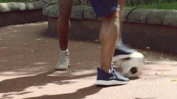 A man is seen gearing up for a soccer match in a public park. He stands beside a black and white soccer ball on a well-maintained, sunny path surrounded by greenery. video
