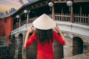 back of girl traveler tourist in Vietnamese hat at ancient Japanese covered bridge in old town in Hoi An in Vietnam photo