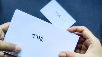 Close up view of a child's hand holding a white envelope labeled THR or Eid Allowance written on it on a black background. This envelope is usually given to family and relatives before Eid al-Fitr. photo