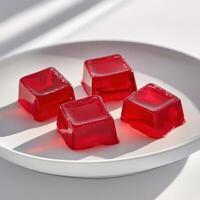 Four vibrant red jelly cubes glisten on a white plate under soft natural light. photo