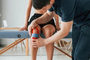 Working with legs. Woman visiting physiologist that putting kinesio tape on the skin, photo