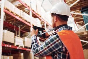 Holding tablet in hands. Employee in uniform is working in the storage at daytime photo