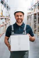 Holding white box and smiling, Employee in uniform is working in the storage at daytime,, photo