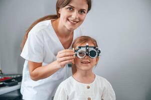 Looking forward, correcting eye sight by device. Little girl have their vision checking in the clinic by help of the female doctor photo
