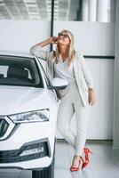 Leaning on the automobile. ,Woman in white formal clothes is in the car dealership photo