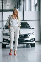 Standing, and posing. Woman in white formal clothes is in the car dealership photo
