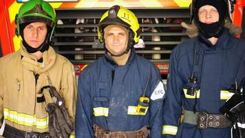 Portrait of male firefighters in helmets and protective uniforms standing near a big red car. Young confident firemen in full equipment posing against the background of a fire engine at station photo