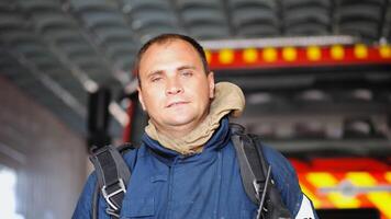 Portrait of tired male firefighter standing near a fire engine at department. Young exhausted fireman in protective uniform looking at camera after fire against the background of big red truck photo