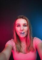 Young woman making a selfie with her tongue out on a dark background. Girl Blowing Bubblegum photo