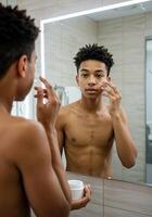 african american man looking at mirror and applying cream on face in bathroom. Man applying face cream photo