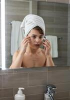 boy applying face mask in front of mirror with towel on his head in bathroom. Boy applying face mask photo