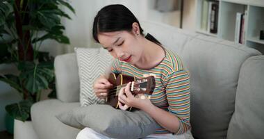 Handheld high angle view shot, Relaxed Asian woman sitting on a sofa enjoying playing the ukulele in a cozy room at home. Practice and dedication to favorite musical instrument. video