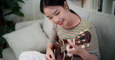 Handheld high angle view shot, Cheerful Asian woman sitting on a sofa enjoying playing the ukulele in a cozy room at home. Practice and dedication to favorite musical instrument. video