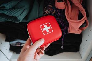 Compact Red First Aid Kit Held by Hand Amongst Clothing in a Neatly Organized Closet photo