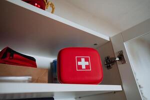 Compact Red First Aid Kit strategically placed on a shelf A collection of essential emergency supplies designed for both home use and travel convenience and safety photo