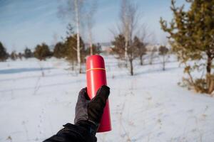 A person is bundled up in warm and cozy winter clothing, standing in the midst of pristine white snow, while holding a bright red thermos that starkly contrasts with the icy white backdrop photo