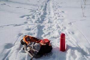 A colorful backpack, paired with a bright red thermos, is resting in the pristine white snow, producing a striking contrast to the beautiful winter landscape surrounding it all photo
